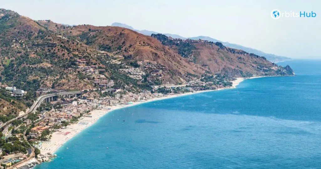 A scenic view of a coastal town along the Ionian Sea, nestled at the base of rugged mountains with clear blue waters extending into the horizon